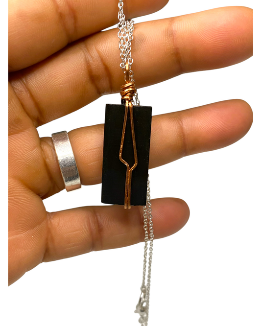 Hand holding a necklace with a black pendant and copper wire design on a white background