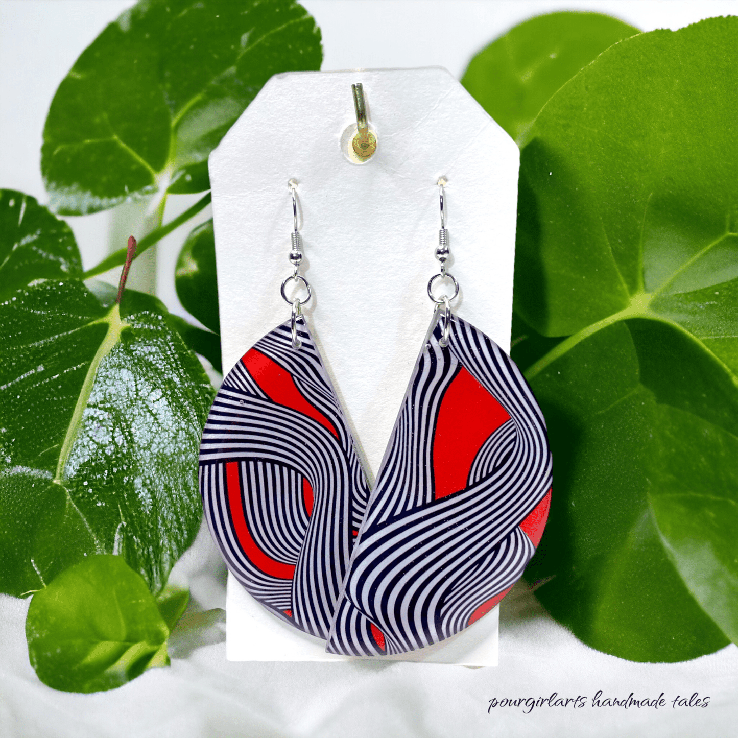 Pair of earrings with red and black wave pattern on a white card with green leaves in the background